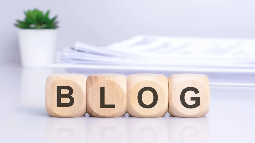 Quattro cubi di legno naturale disposti in fila che formano la parola "BLOG". Ogni cubo presenta una lettera nera stampata chiaramente sulla superficie frontale (B-L-O-G).